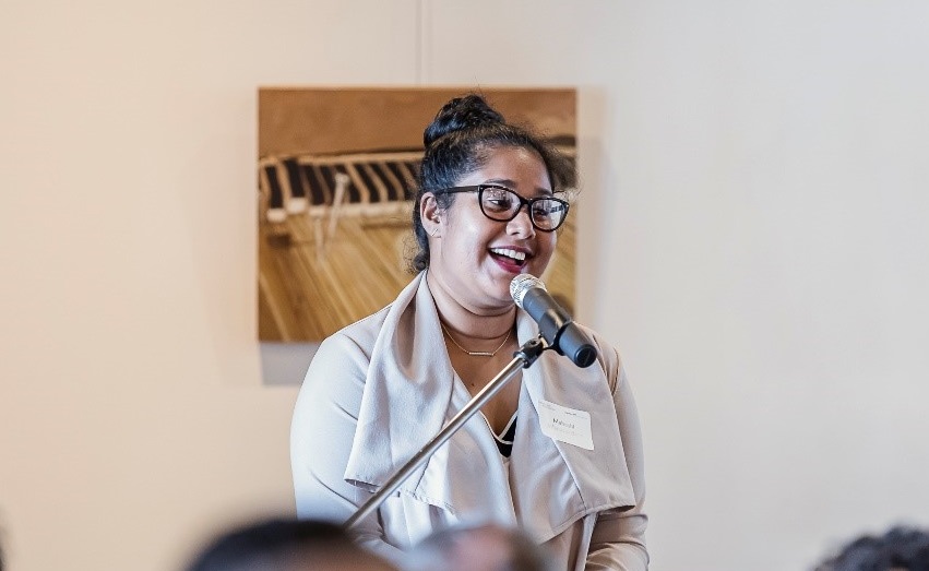 Smiling woman of colour in glasses and a khaki dress in front of a microphone addressing a room of people